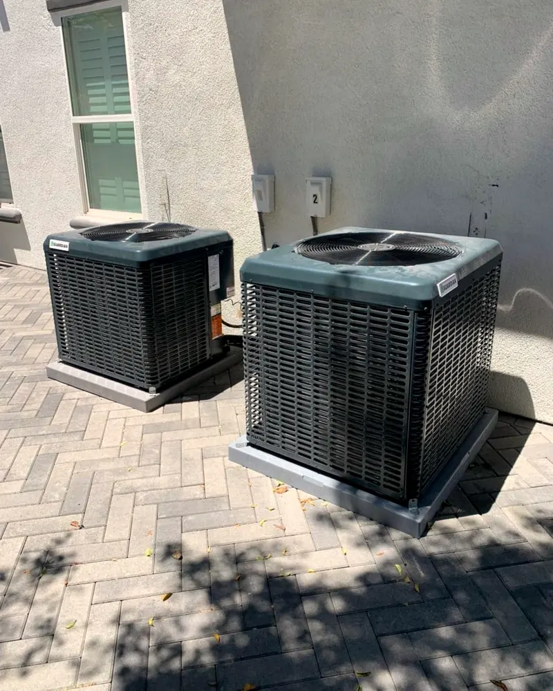 Two new outdoor AC units side by side on a paver patio next to a home